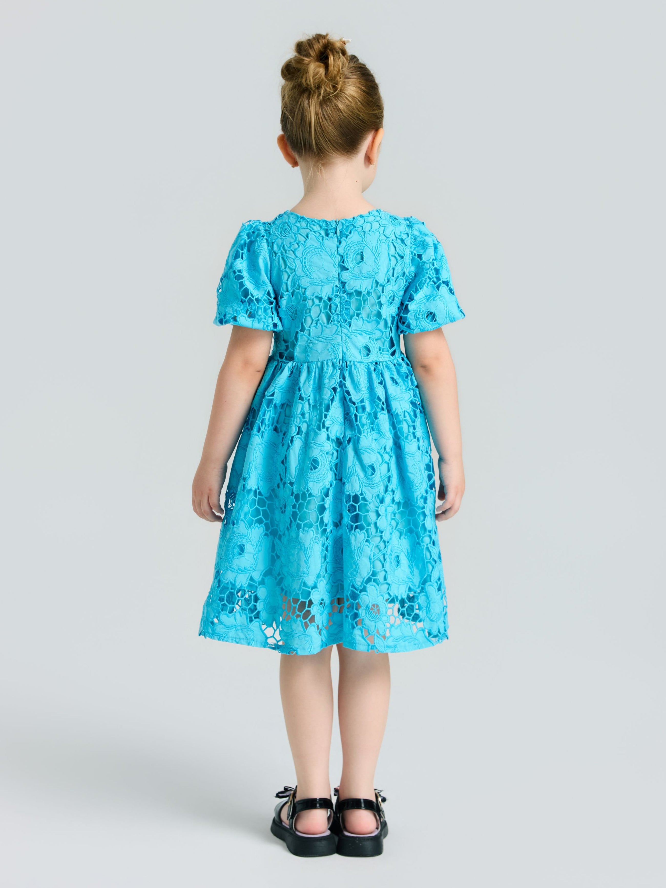 A young girl faces away in the Social Queen Girls 3D Floral Appliqué Cutwork Lace Dress featuring short sleeves and black shoes, set against a light gray background.