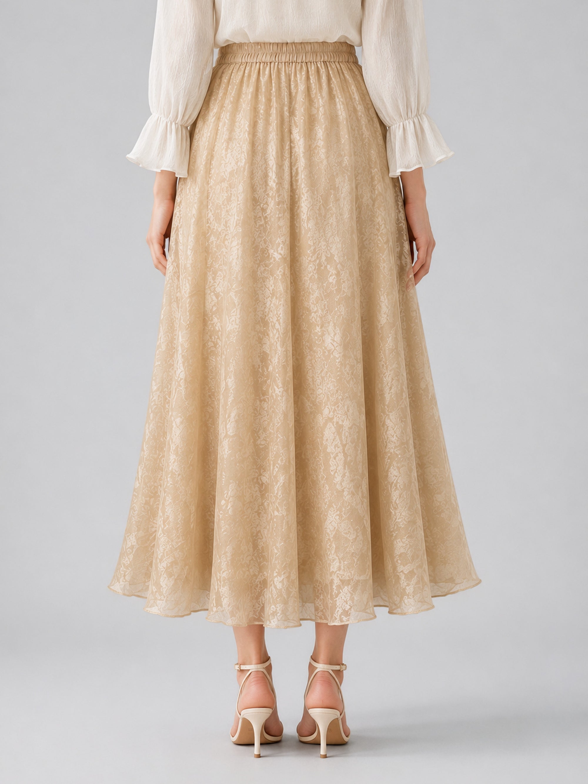 A person stands facing away, wearing a cream blouse with ruffled sleeves and the Social Queen Jacquard Chiffon Midi Skirt, featuring a beige jacquard texture and elastic waist, elegantly paired with light strappy heels.