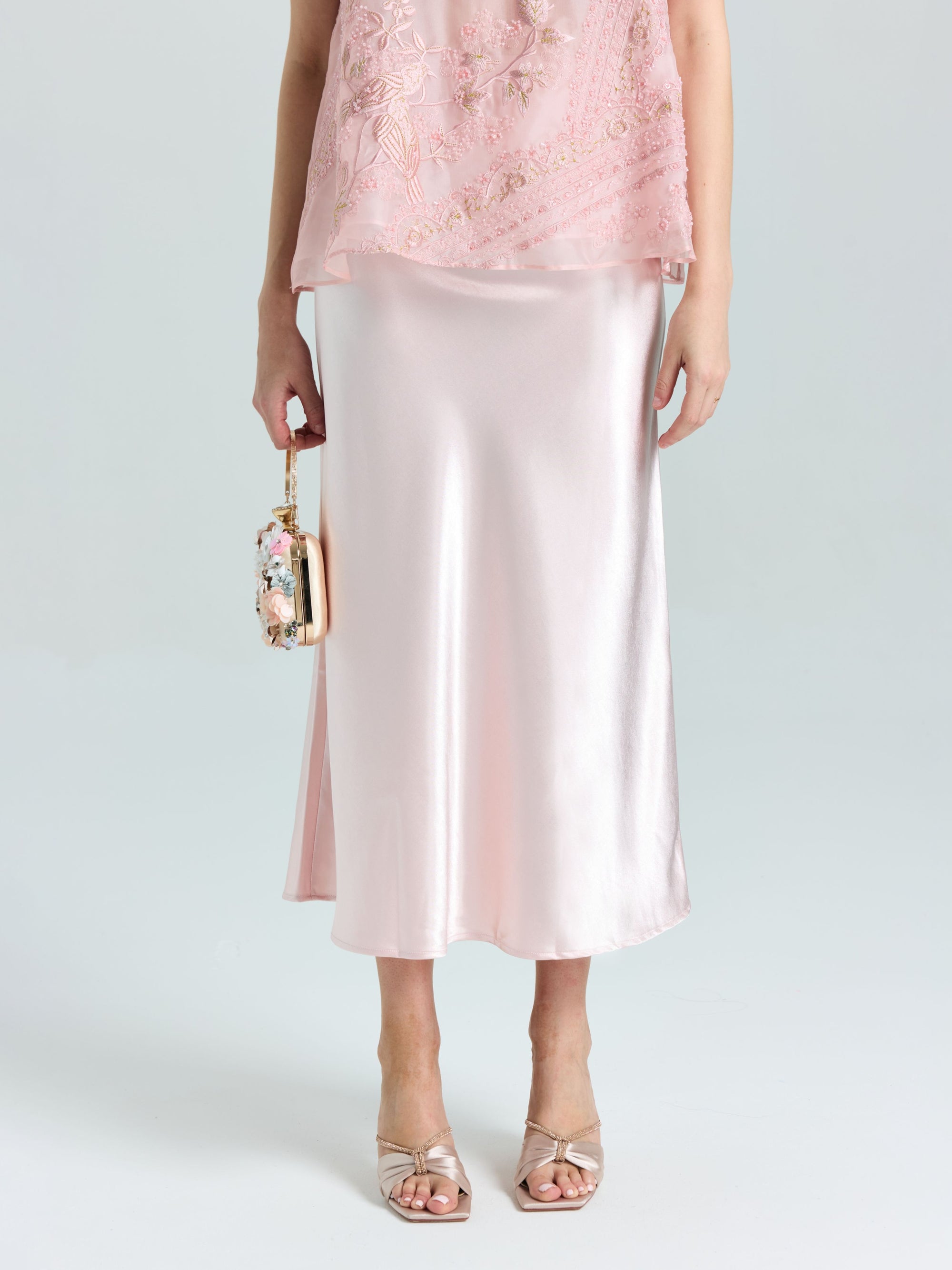 A person wears the Social Queen Satin A-Line Midi Skirt in pale pink with an embellished top, open-toe heels, and holds a small beaded handbag.