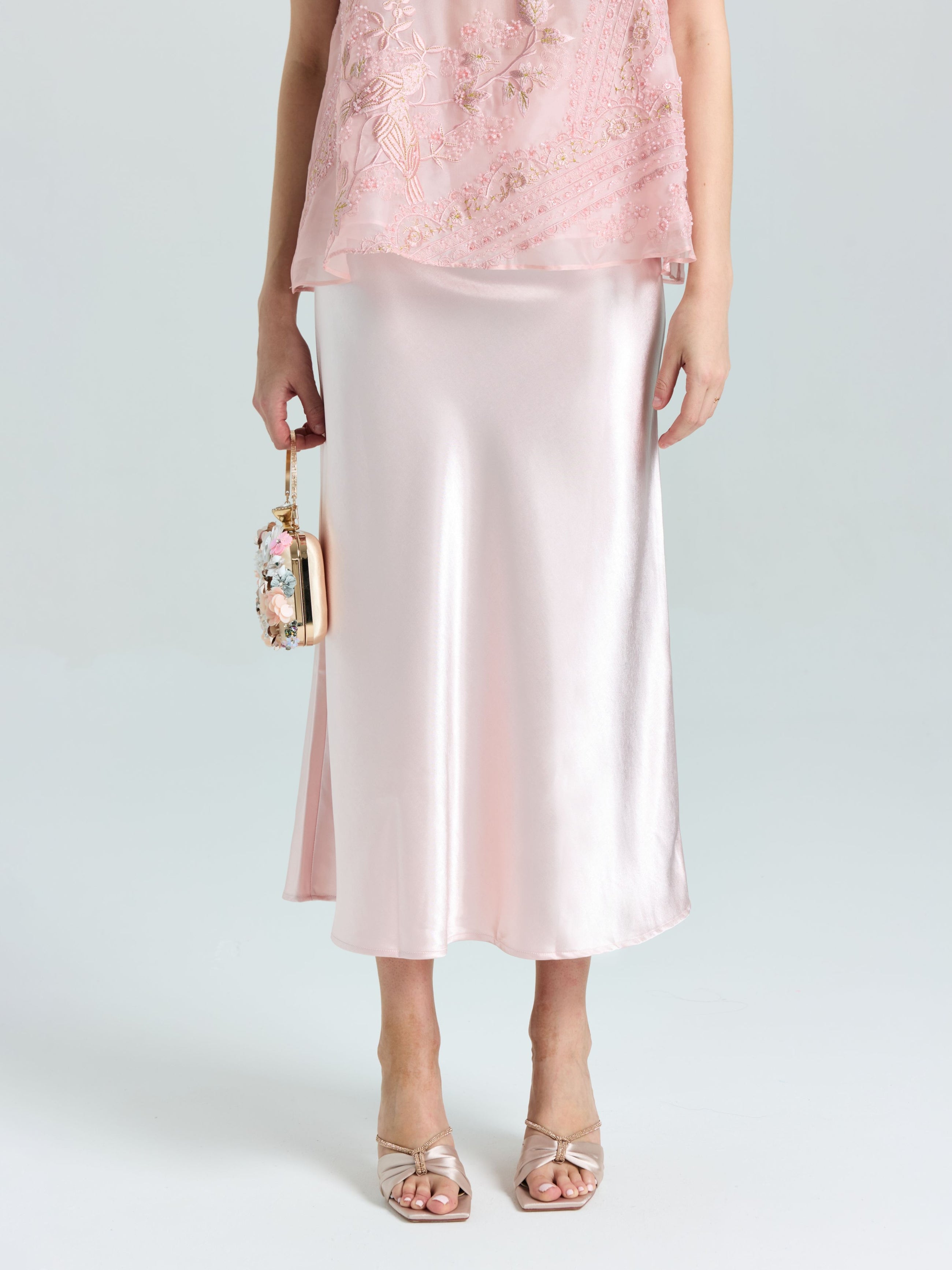 A person wears the Social Queen Satin A-Line Midi Skirt in pale pink with an embellished top, open-toe heels, and holds a small beaded handbag.