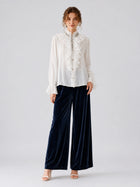 A person stands against a plain background wearing the Social Queen Pearl Embellished Ruffle Collar Blouse, navy blue wide-leg velvet pants, and white heeled sandals.