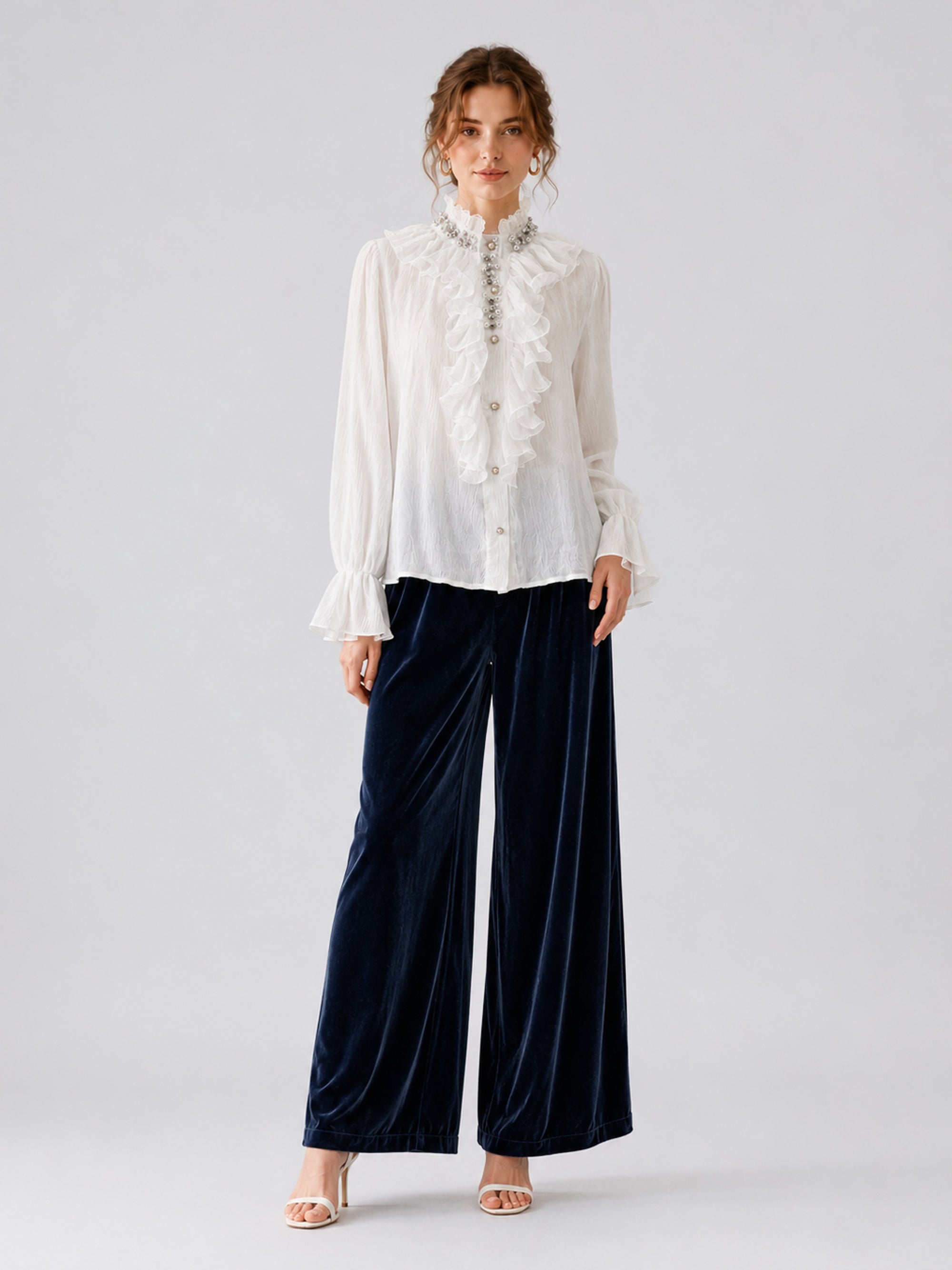 A person stands against a plain background wearing the Social Queen Pearl Embellished Ruffle Collar Blouse, navy blue wide-leg velvet pants, and white heeled sandals.