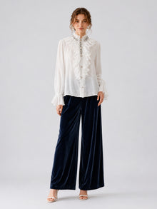 A person stands against a plain background wearing the Social Queen Pearl Embellished Ruffle Collar Blouse, navy blue wide-leg velvet pants, and white heeled sandals.