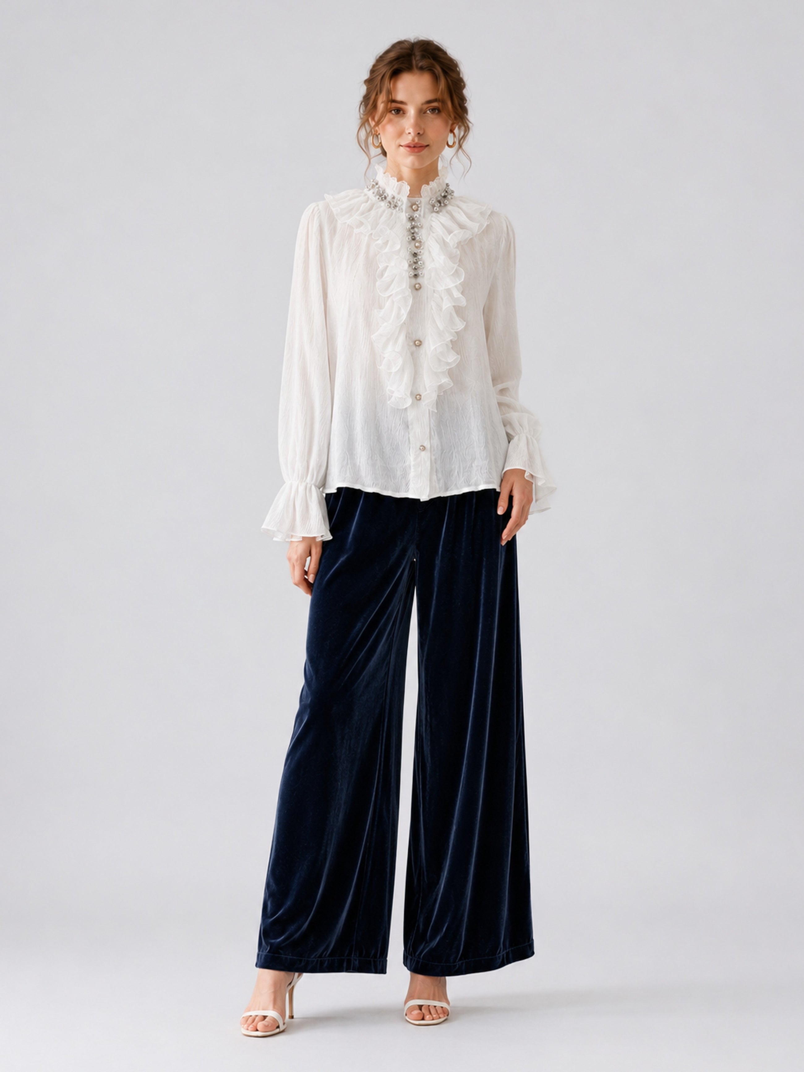 A person stands against a plain background wearing the Social Queen Pearl Embellished Ruffle Collar Blouse, navy blue wide-leg velvet pants, and white heeled sandals.
