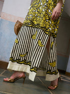 Wearing the Social Queen Tropical Fruit Print Linen Dress, which features a long silhouette with vibrant yellow flowers and bananas reminiscent of airy resort elegance, this person stands on a tiled floor paired with gold heeled sandals and holding a wooden bag.