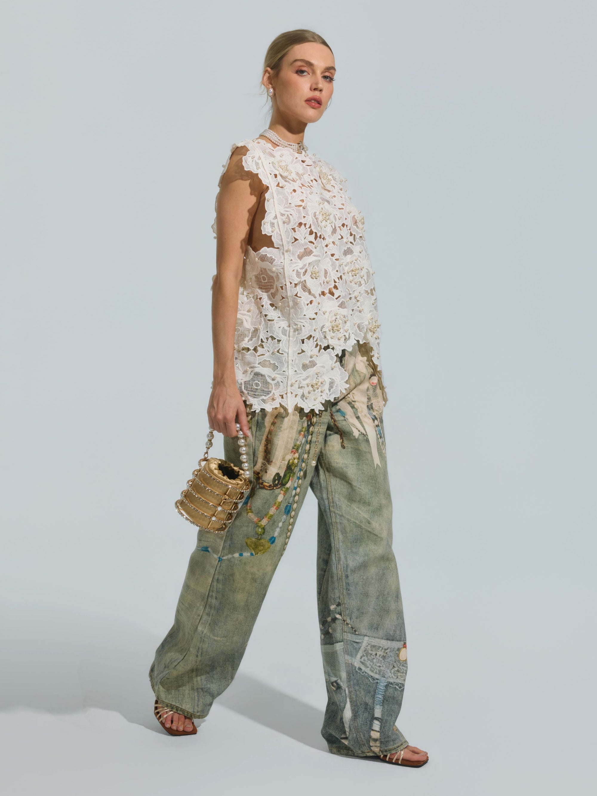 A woman wearing the Social Queen Pearl Rose Guipure Lace Top, paired with loose patchwork denim jeans and sandals, holds a small gold handbag with pearl details while posing against a plain light background.