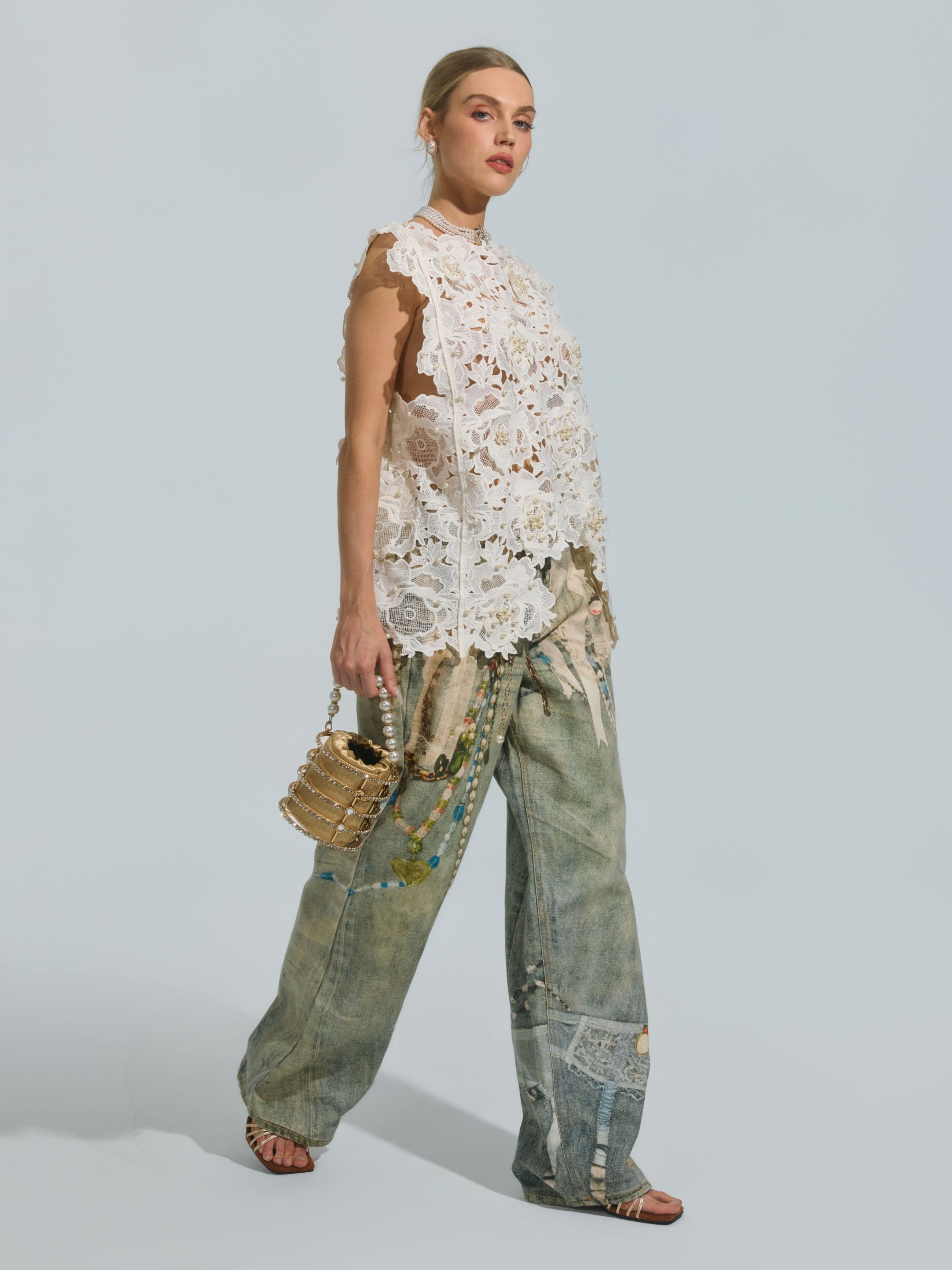 A woman wearing the Social Queen Pearl Rose Guipure Lace Top, paired with loose patchwork denim jeans and sandals, holds a small gold handbag with pearl details while posing against a plain light background.