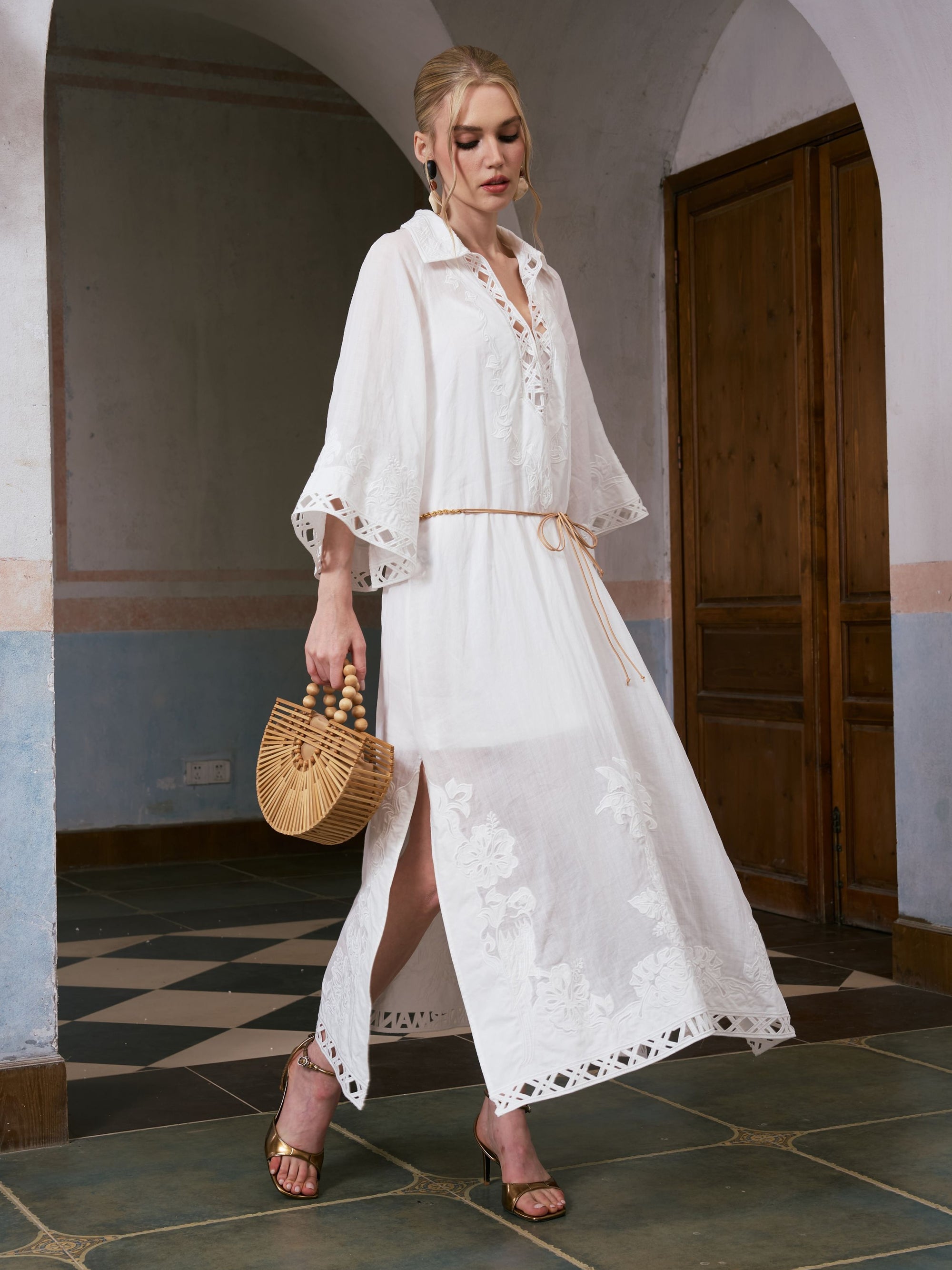 A woman walks indoors on a tiled floor wearing the Social Queen Floral Cutwork Embroidered Linen Dress—a long white linen maxi dress featuring lace details and delicate floral cutwork embroidery—paired with gold sandals and carrying a small wooden handbag.