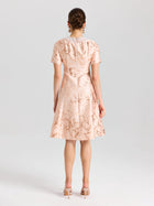 A woman faces away from the camera, wearing Social Queen's Pearl Embellished Jacquard Dress—a short-sleeved, knee-length satin piece in light pink floral print—with heeled sandals, set against a plain background.