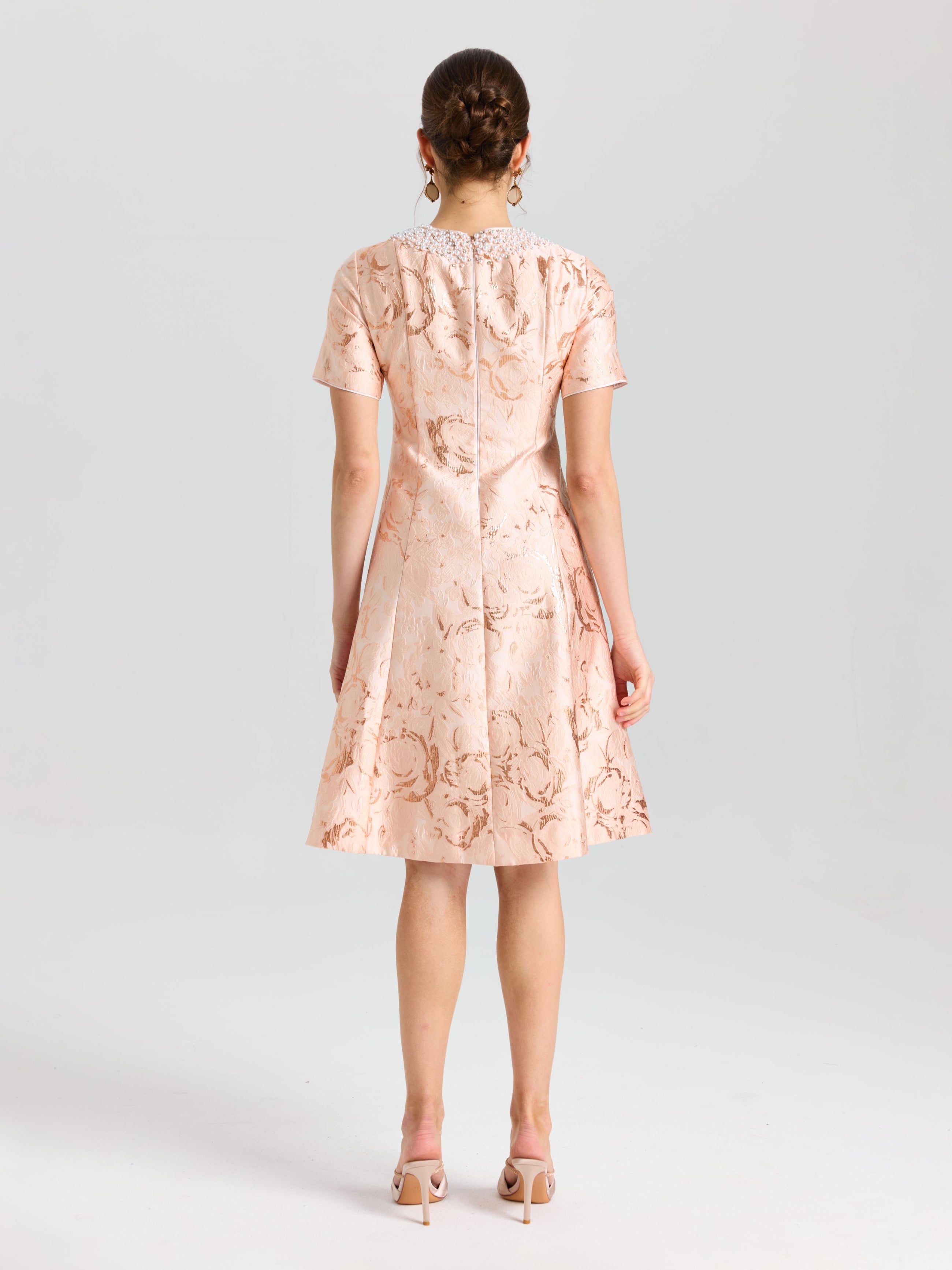 A woman faces away from the camera, wearing Social Queen's Pearl Embellished Jacquard Dress—a short-sleeved, knee-length satin piece in light pink floral print—with heeled sandals, set against a plain background.