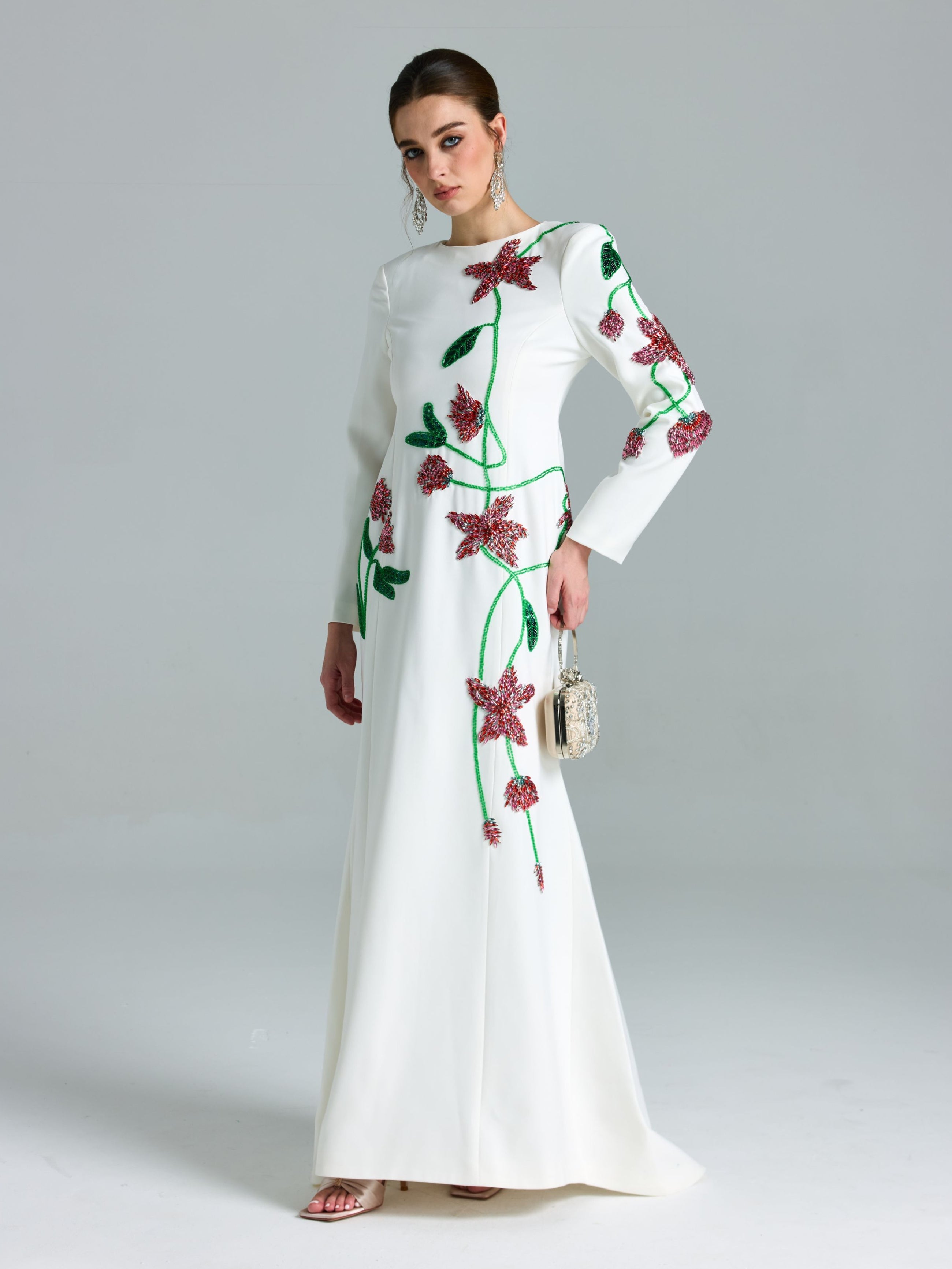 A woman poses in the Social Queen Floral Beaded A-Line Gown, a long-sleeved white occasionwear dress with red floral patterns, and carries a small silver handbag against a plain backdrop.