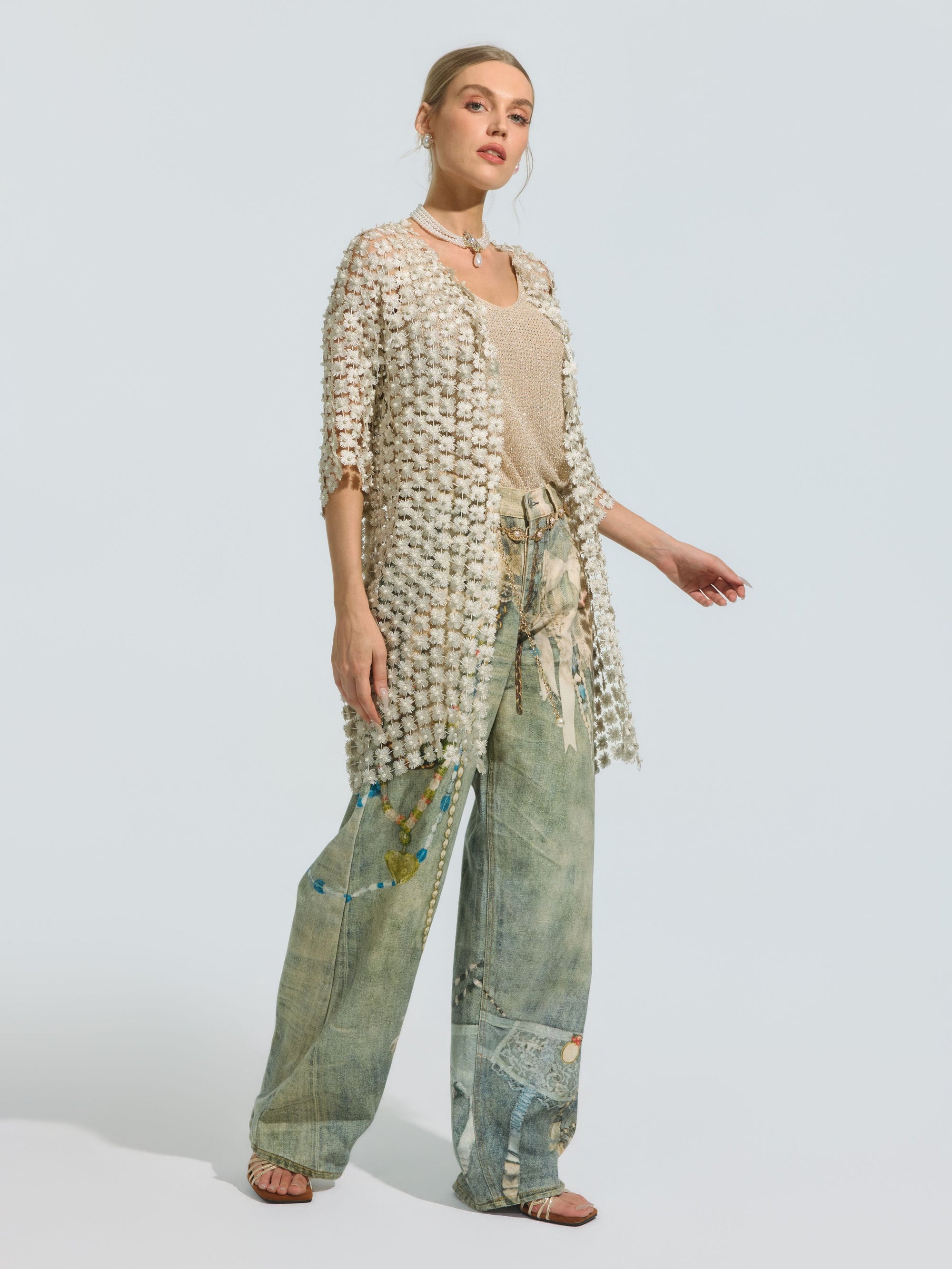 A woman wears the Social Queen Pearl Daisy Cutwork Long Cardigan, a sheer and lightweight polyester piece (camisole not included), styled over a beige top and wide-leg, patterned patchwork denim jeans. She completes the look with sandals and layered pearl-detailed necklaces.