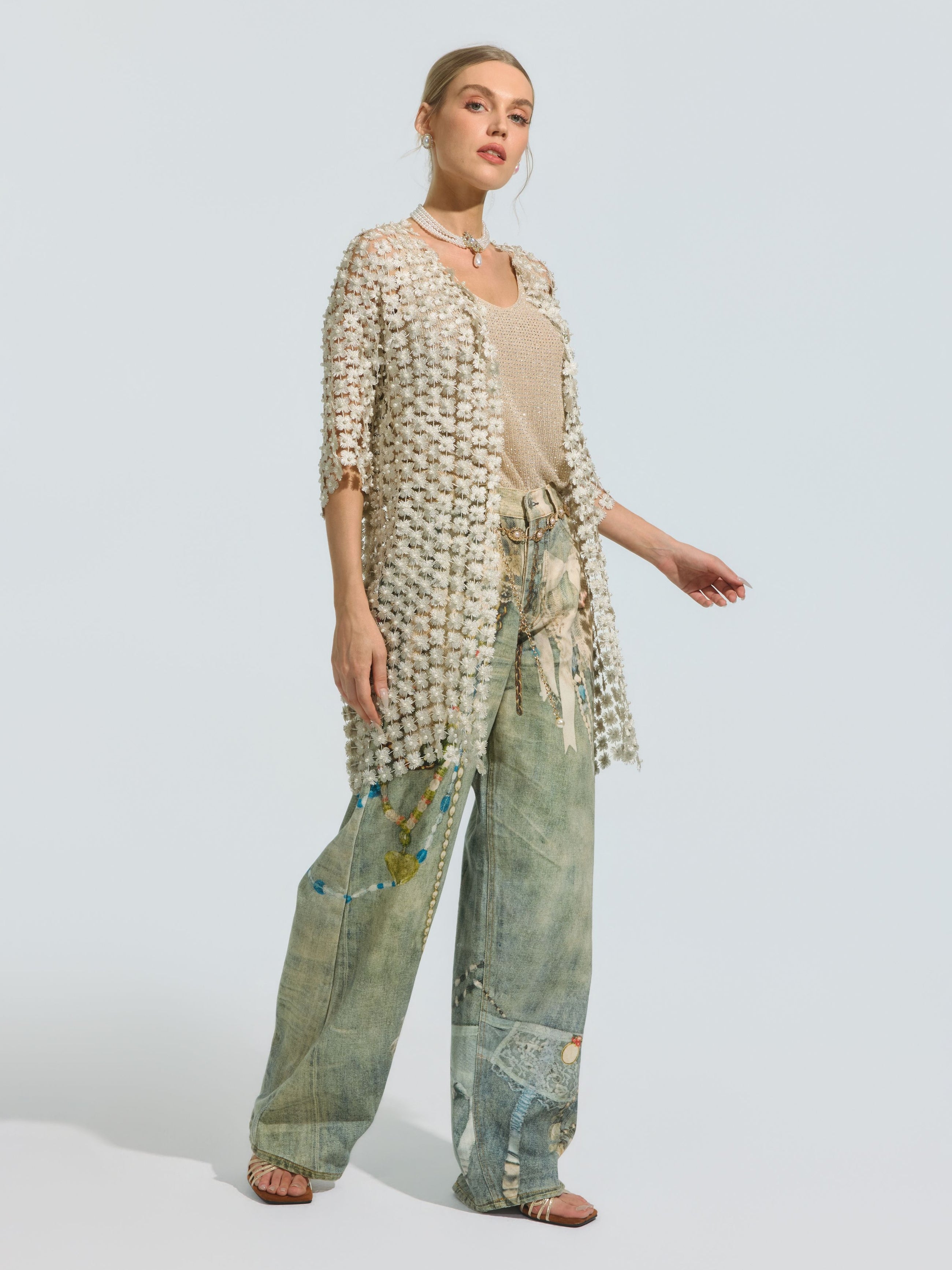 A woman wears the Social Queen Pearl Daisy Cutwork Long Cardigan, a sheer and lightweight polyester piece (camisole not included), styled over a beige top and wide-leg, patterned patchwork denim jeans. She completes the look with sandals and layered pearl-detailed necklaces.