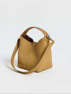 The Social Queen Woven Handle Pebbled Leather Bucket Bag, featuring a light brown pebbled finish, woven top handle, and detachable shoulder strap, is shown against a plain white background.