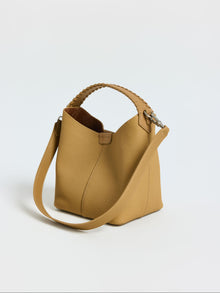The Social Queen Woven Handle Pebbled Leather Bucket Bag, featuring a light brown pebbled finish, woven top handle, and detachable shoulder strap, is shown against a plain white background.