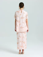 A woman stands facing away, wearing the Social Queen Dragonfly Beaded Lace Top in light pink with delicate embroidery and a matching long skirt. Her hair is up and she completes the look with nude heels.