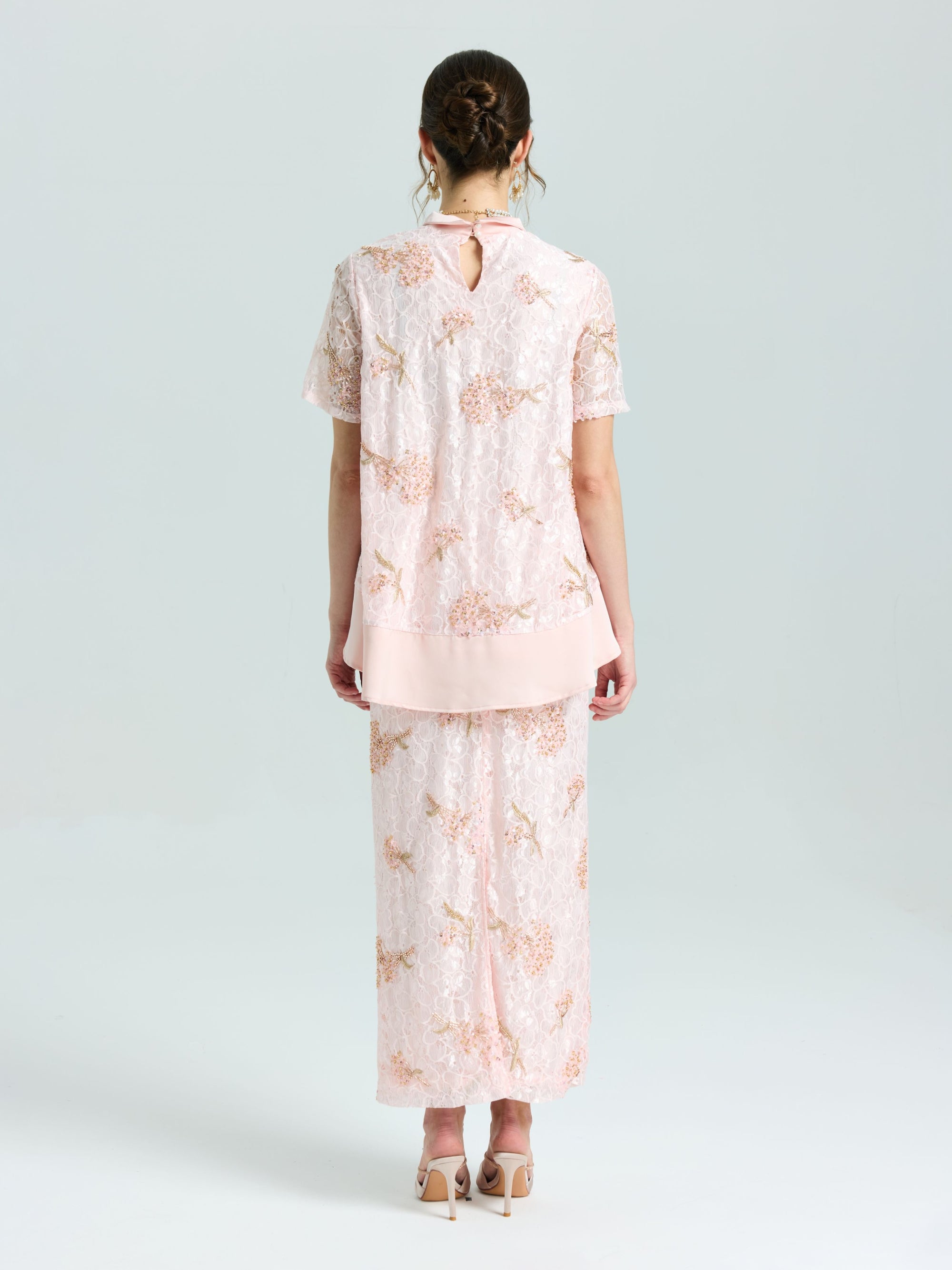 A woman stands facing away, wearing the Social Queen Dragonfly Beaded Lace Top in light pink with delicate embroidery and a matching long skirt. Her hair is up and she completes the look with nude heels.