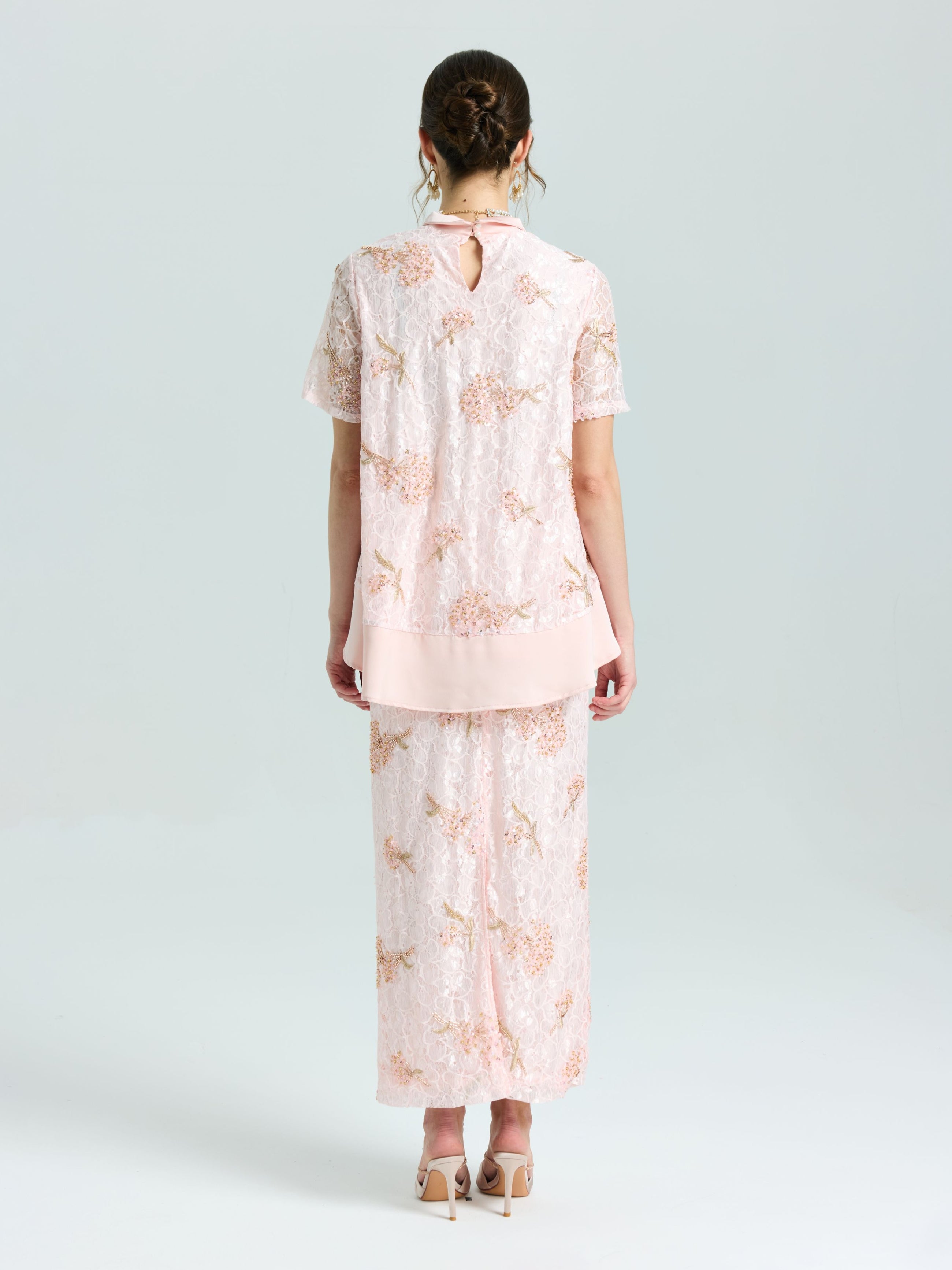 A woman stands facing away, wearing the Social Queen Dragonfly Beaded Lace Top in light pink with delicate embroidery and a matching long skirt. Her hair is up and she completes the look with nude heels.