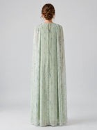 A person stands with their back to the camera, wearing the Social Queen Beaded Printed Cape Chiffon Dress—a long, light green gown featuring a patterned chiffon overlay and sheer fabric.