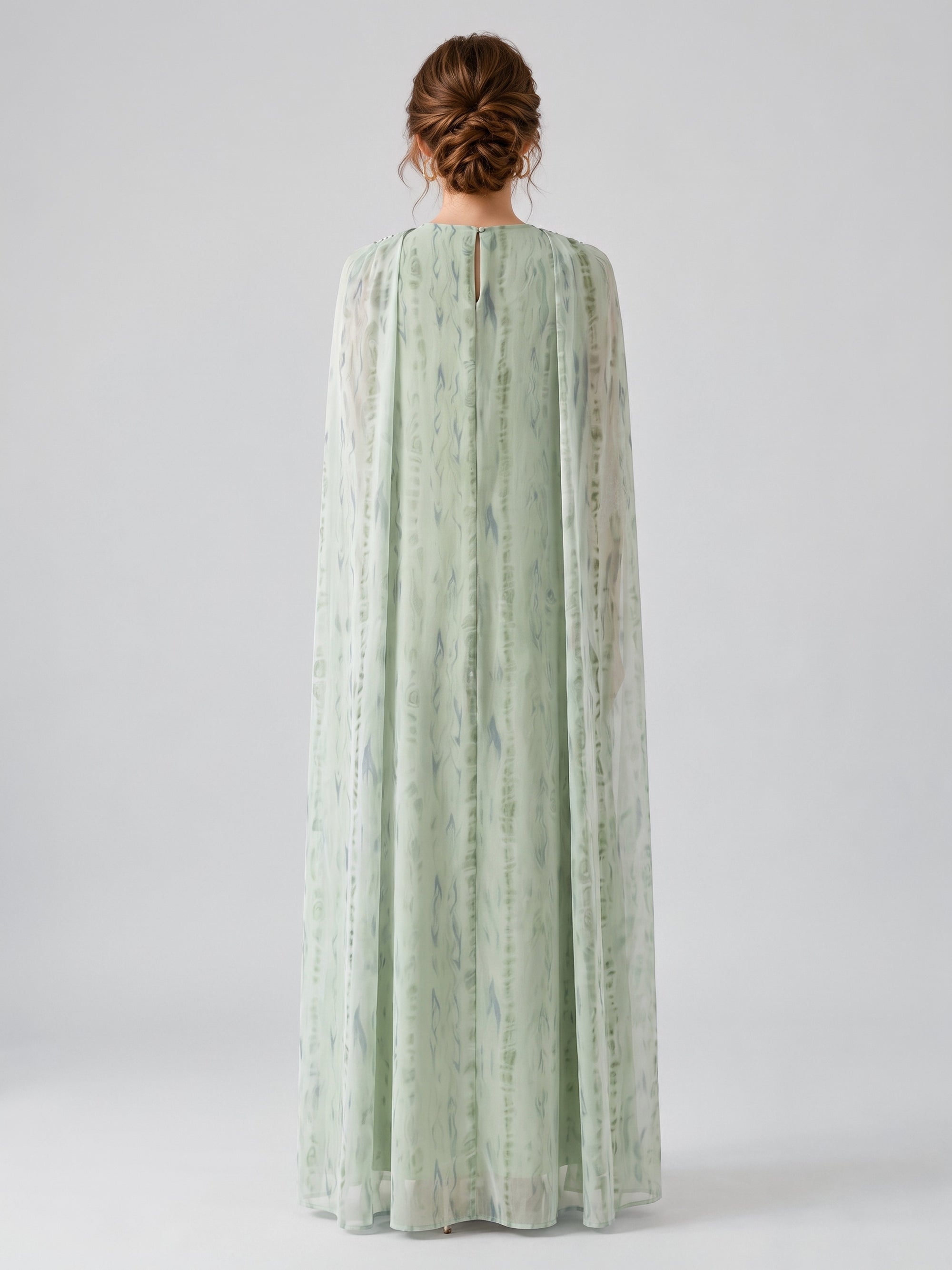 A person stands with their back to the camera, wearing the Social Queen Beaded Printed Cape Chiffon Dress—a long, light green gown featuring a patterned chiffon overlay and sheer fabric.