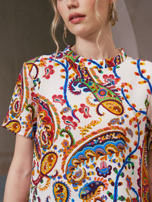 A woman wearing the Social Queen Paisley Garden Print Silk Top, a white short-sleeve shirt adorned with intricate red, blue, yellow, and orange paisley designs—a perfect choice for resort wear.