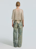 A person stands facing away, wearing the Social Queen Pearl Daisy Cutwork Top (camisole not included), which features a textured grid-patterned design. They pair it with loose, faded jeans embellished with long chains and beads. Their hair is tied in a neat bun, set against a plain light background.