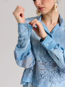 A woman in the Social Queen Washed Silk Lace Detail Shirt, a relaxed fit light blue blouse with lace trim, fastens a cuff button. Only her upper body is visible against a plain background.