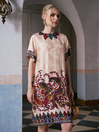 A woman stands indoors wearing the Social Queen Paisley Garden Print Jacquard Silk Dress, featuring a cream-colored satin base with vibrant, intricate patterns adorning the collar, sleeves, and hem. Her blond hair is pulled back as she faces slightly to the right.