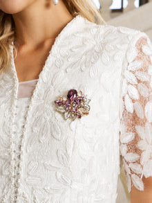 A woman in a white textured, floral-patterned dress with lace sleeves wears the Social Queen Amethyst Floral Brooch—an oversized gold-tone piece accented with amethyst crystal—pinned to her chest.