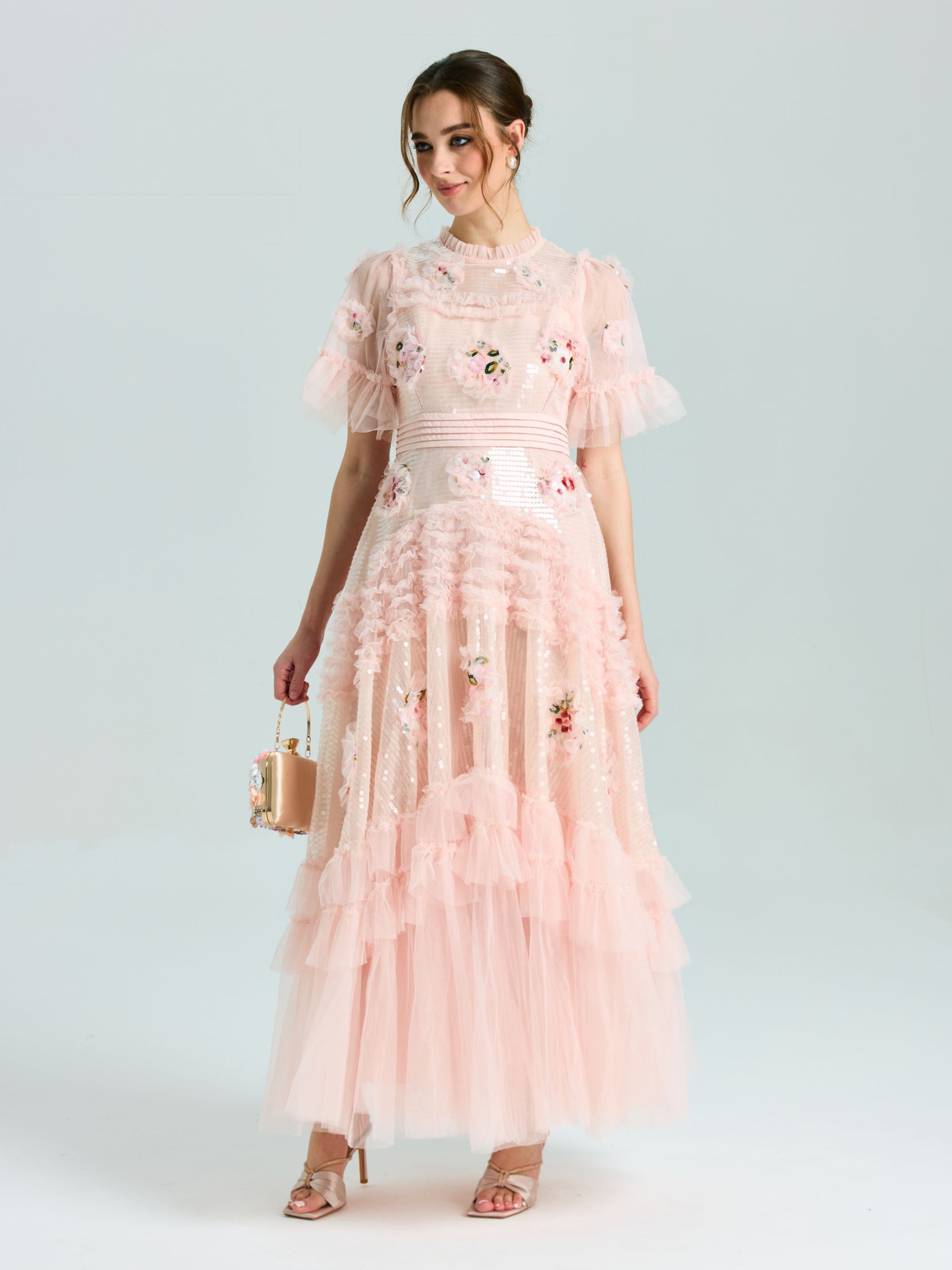 A woman wears the Social Queen Embroidered Ruffle Tiered Tulle Dress in light pink, featuring floral embroidery and a tiered skirt, holding a small handbag against a plain background.