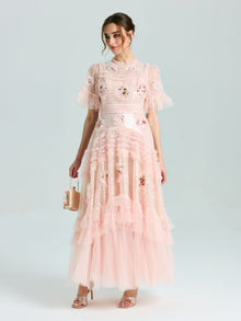 A woman wears the Social Queen Embroidered Ruffle Tiered Tulle Dress in light pink, featuring floral embroidery and a tiered skirt, holding a small handbag against a plain background.