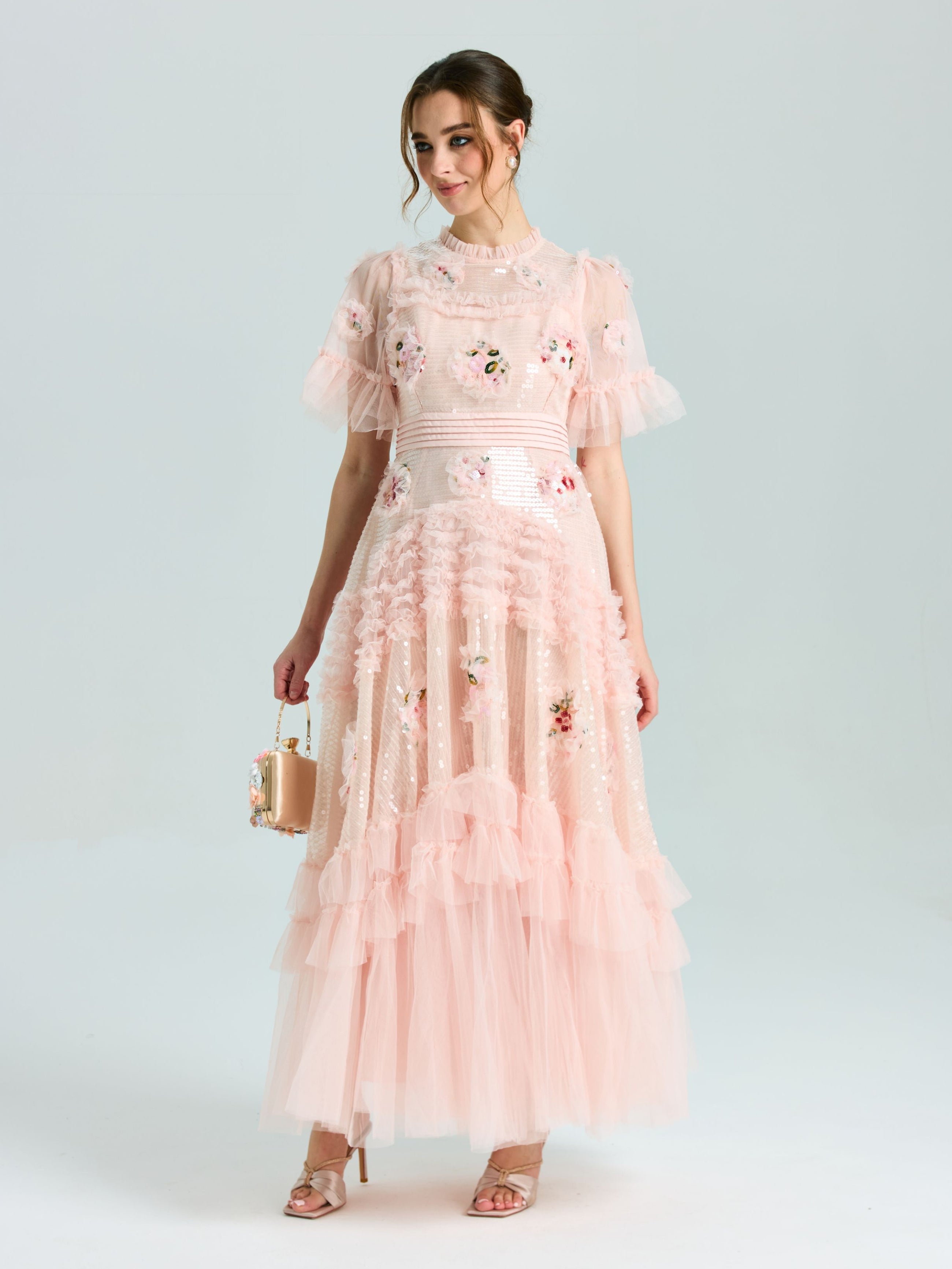 A woman wears the Social Queen Embroidered Ruffle Tiered Tulle Dress in light pink, featuring floral embroidery and a tiered skirt, holding a small handbag against a plain background.