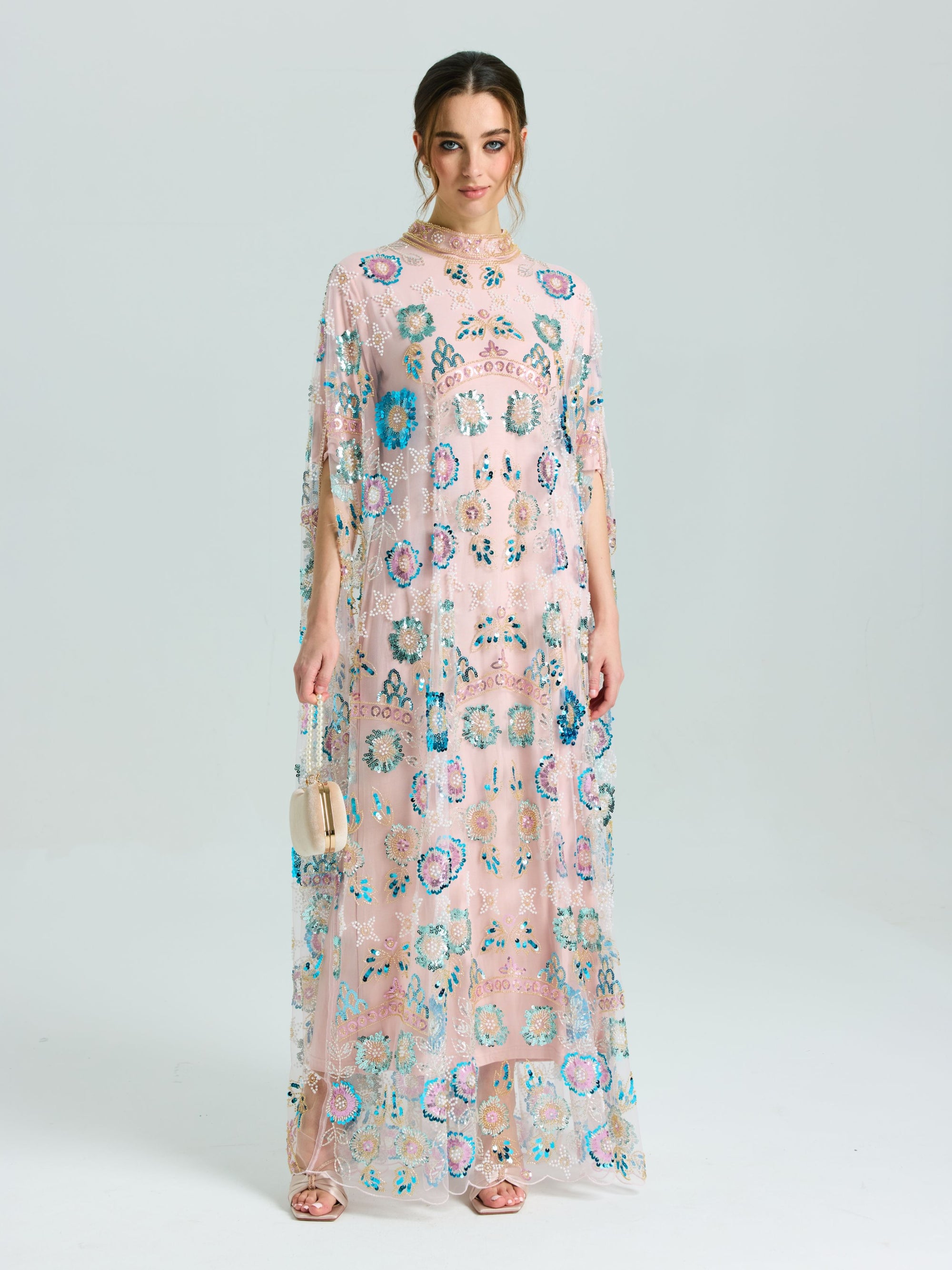 A woman stands against a plain light background wearing Social Queen's Beaded Floral Embellished Cape Gown in light pink with colorful sequins and a beaded neckline, paired with a small beige handbag.