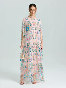 A woman stands against a plain light background wearing Social Queen's Beaded Floral Embellished Cape Gown in light pink with colorful sequins and a beaded neckline, paired with a small beige handbag.