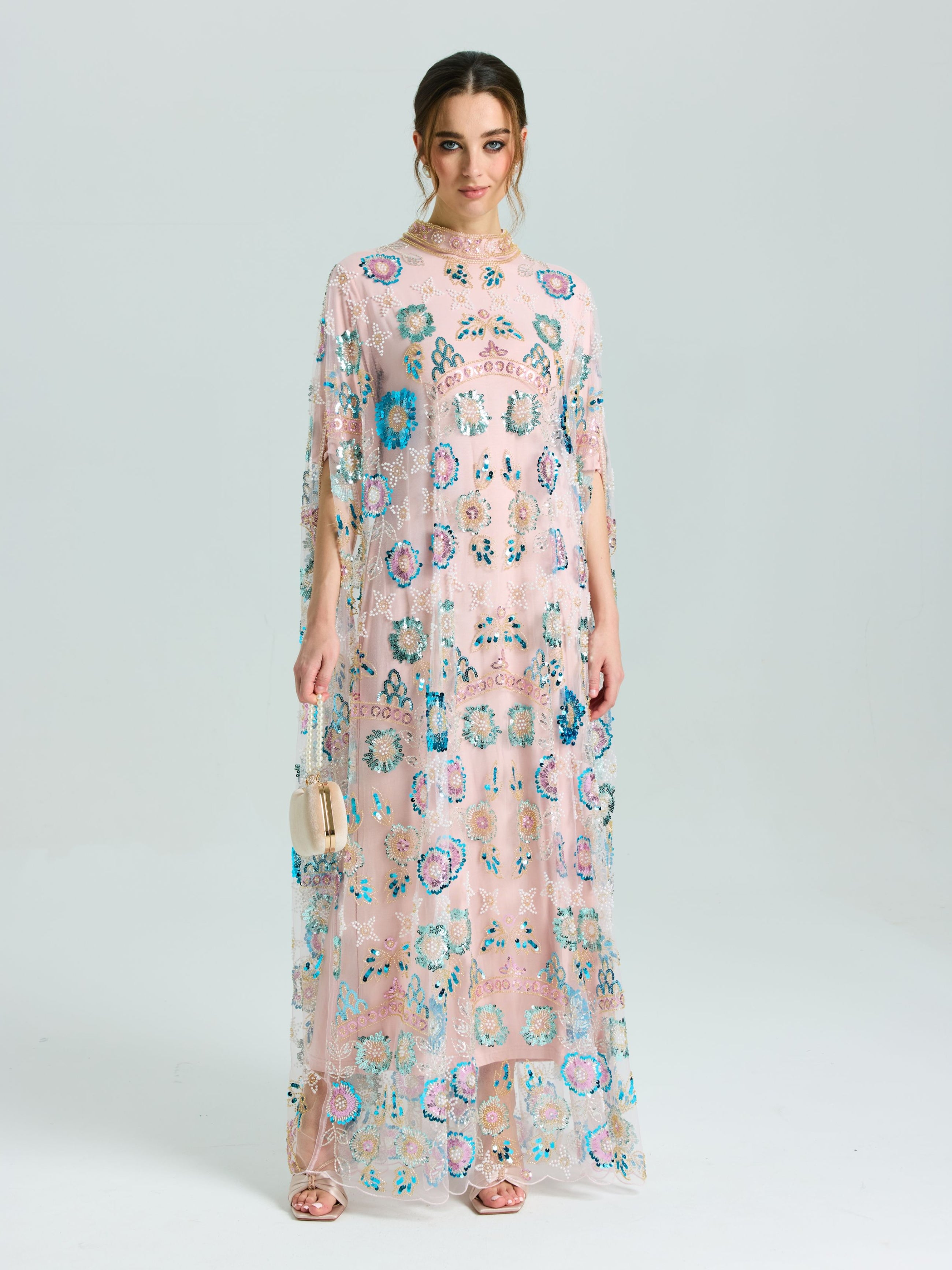 A woman stands against a plain light background wearing Social Queen's Beaded Floral Embellished Cape Gown in light pink with colorful sequins and a beaded neckline, paired with a small beige handbag.