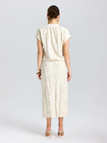 A woman faces away from the camera, wearing Social Queen's Floral Embroidered Linen Midi Skirt in cream with a subtle pattern, paired with a matching short-sleeved top and nude heels.