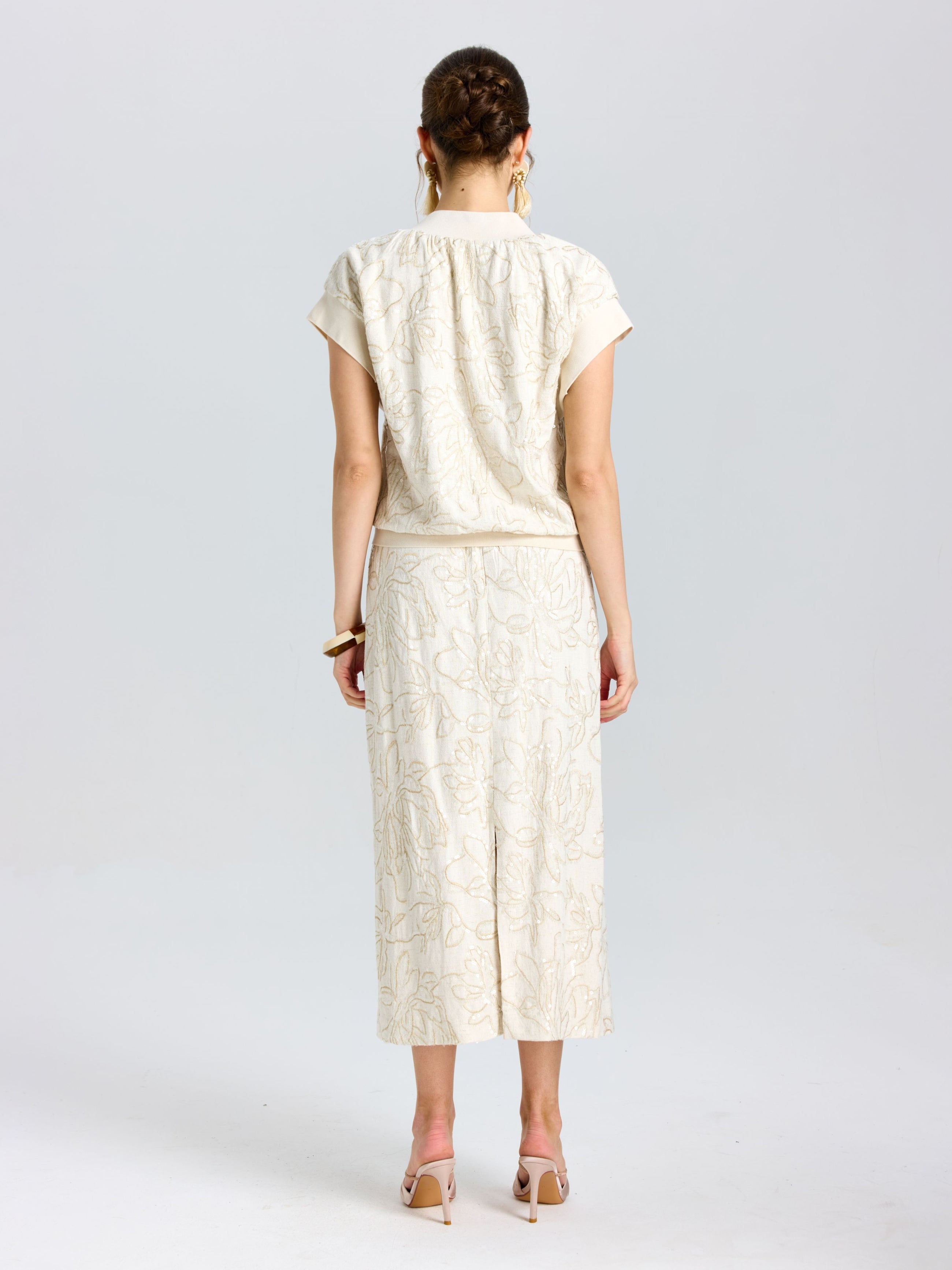 A woman faces away from the camera, wearing Social Queen's Floral Embroidered Linen Midi Skirt in cream with a subtle pattern, paired with a matching short-sleeved top and nude heels.