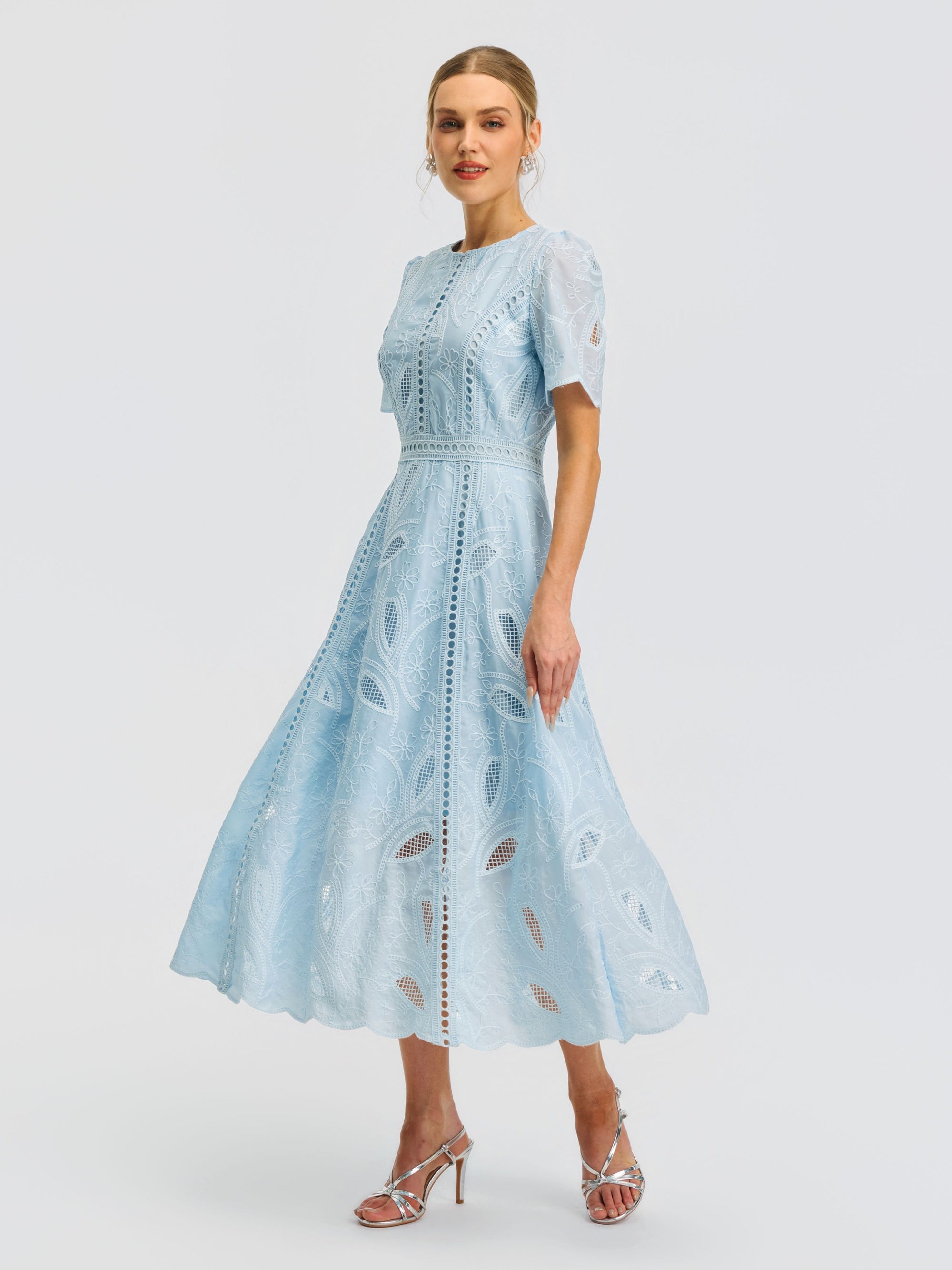 A woman is wearing the Social Queen Floral Cutwork Embroidered Dress, a light blue midi silhouette with lace detailing and short sleeves, paired with silver high-heeled sandals and standing against a plain white background.