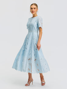 A woman is wearing the Social Queen Floral Cutwork Embroidered Dress, a light blue midi silhouette with lace detailing and short sleeves, paired with silver high-heeled sandals and standing against a plain white background.