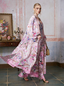 Clad in the Social Queen Baroque Print Mulberry Silk Robe with Scarf, a woman stands indoors, exuding refined elegance. She holds a small gold handbag, while behind her, a painting, candles, and decorative accents on a wooden table set the scene—highlighting the luxurious appeal of this robe as perfect attire for sophisticated gatherings.