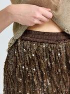 A person lifts a gold mesh top, revealing the Social Queen Sequin Fringe Wide-Leg Pants with a brown elastic waistband, brown fringe, and shimmering sequins.