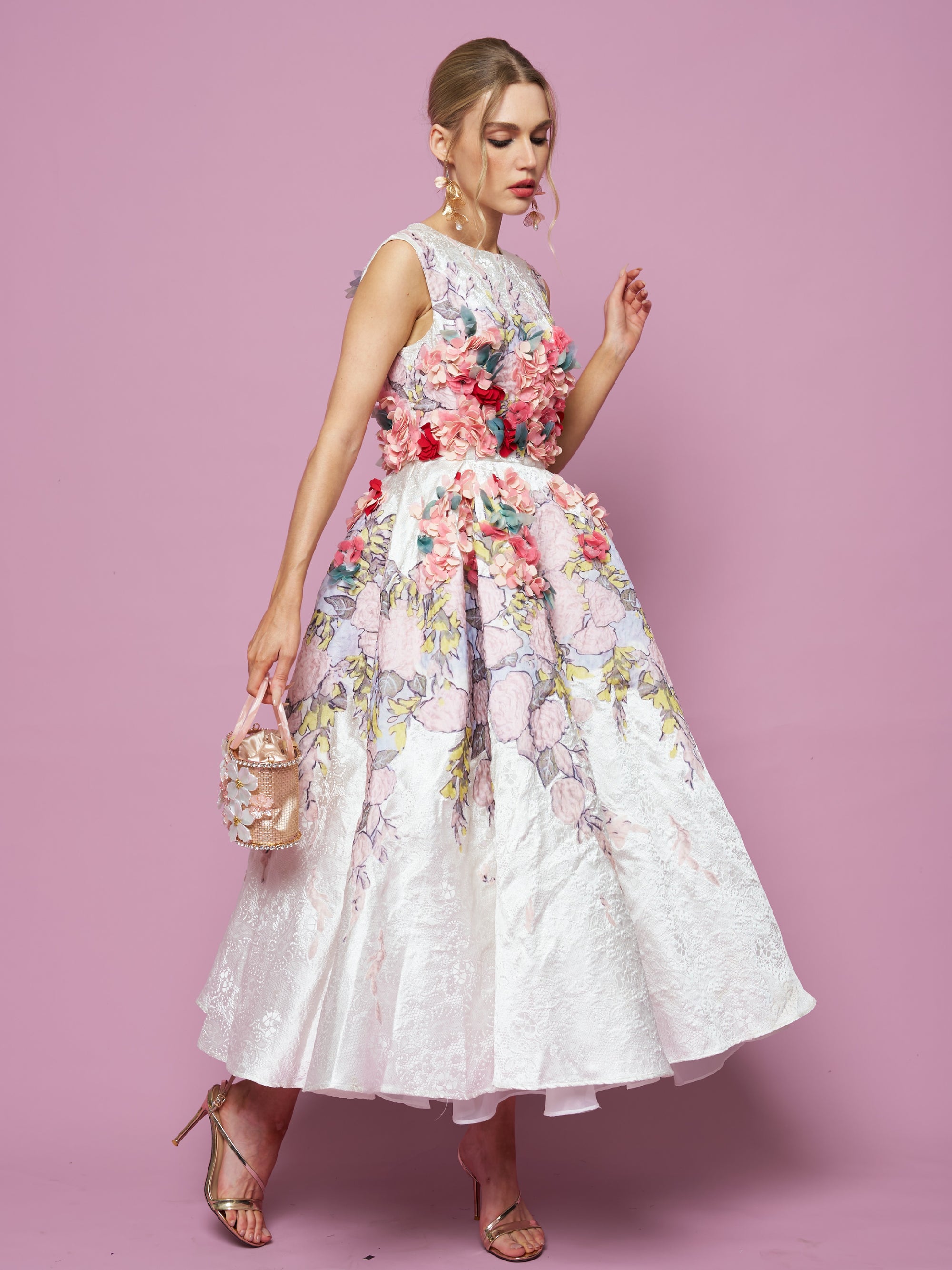 Woman wearing a sleeveless white floral dress with 3D flower embellishments, holding a small purse, and standing against a pink background.