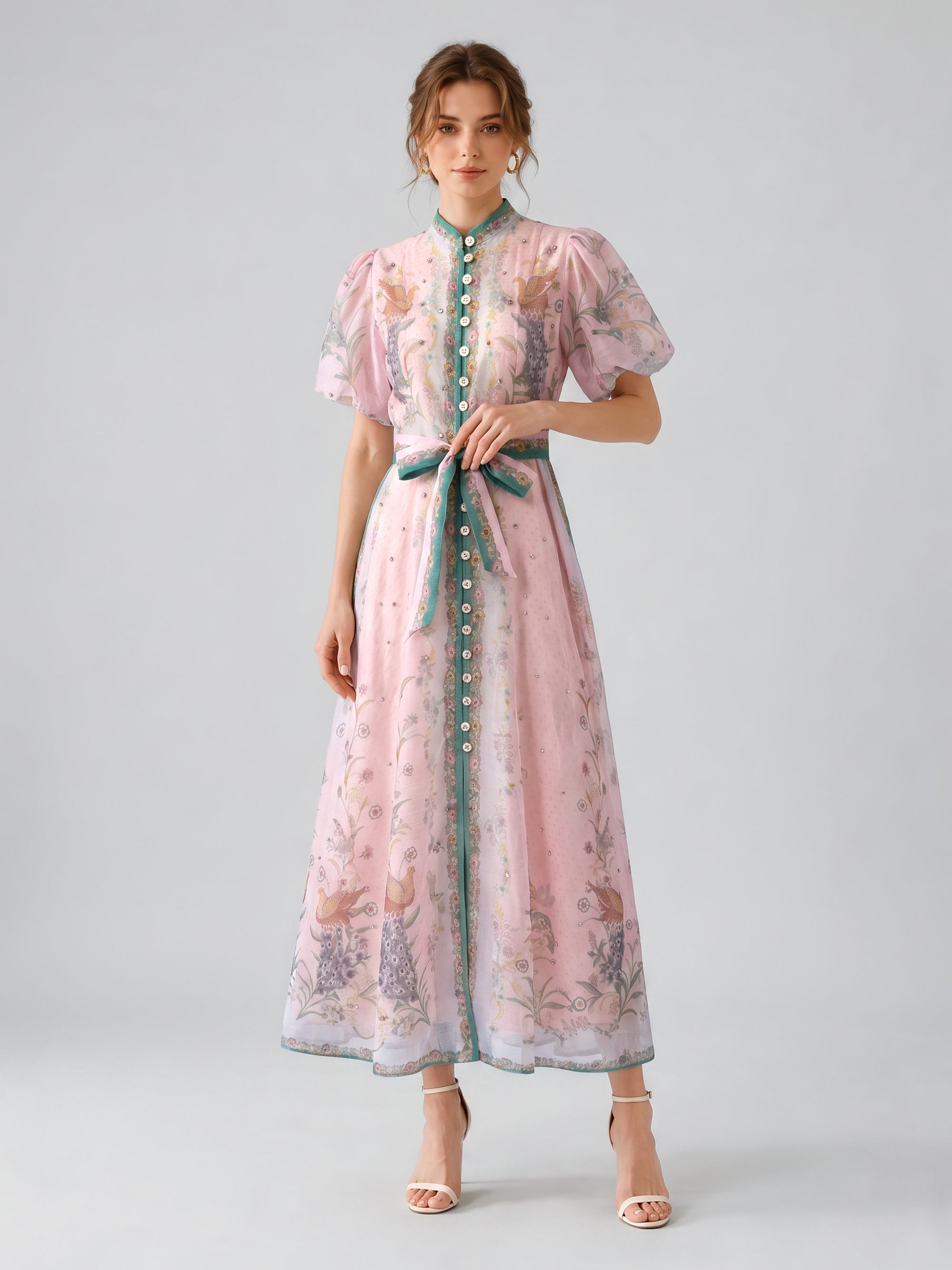 A woman stands against a plain gray background wearing the Social Queen Peacock Botanical Print Dress (Includes Belt), an elegant light pink, ankle-length A-line dress adorned with peacock and floral patterns, green trim, puff sleeves, a bow belt, and white sandals.