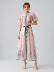 A woman stands against a plain gray background wearing the Social Queen Peacock Botanical Print Dress (Includes Belt), an elegant light pink, ankle-length A-line dress adorned with peacock and floral patterns, green trim, puff sleeves, a bow belt, and white sandals.