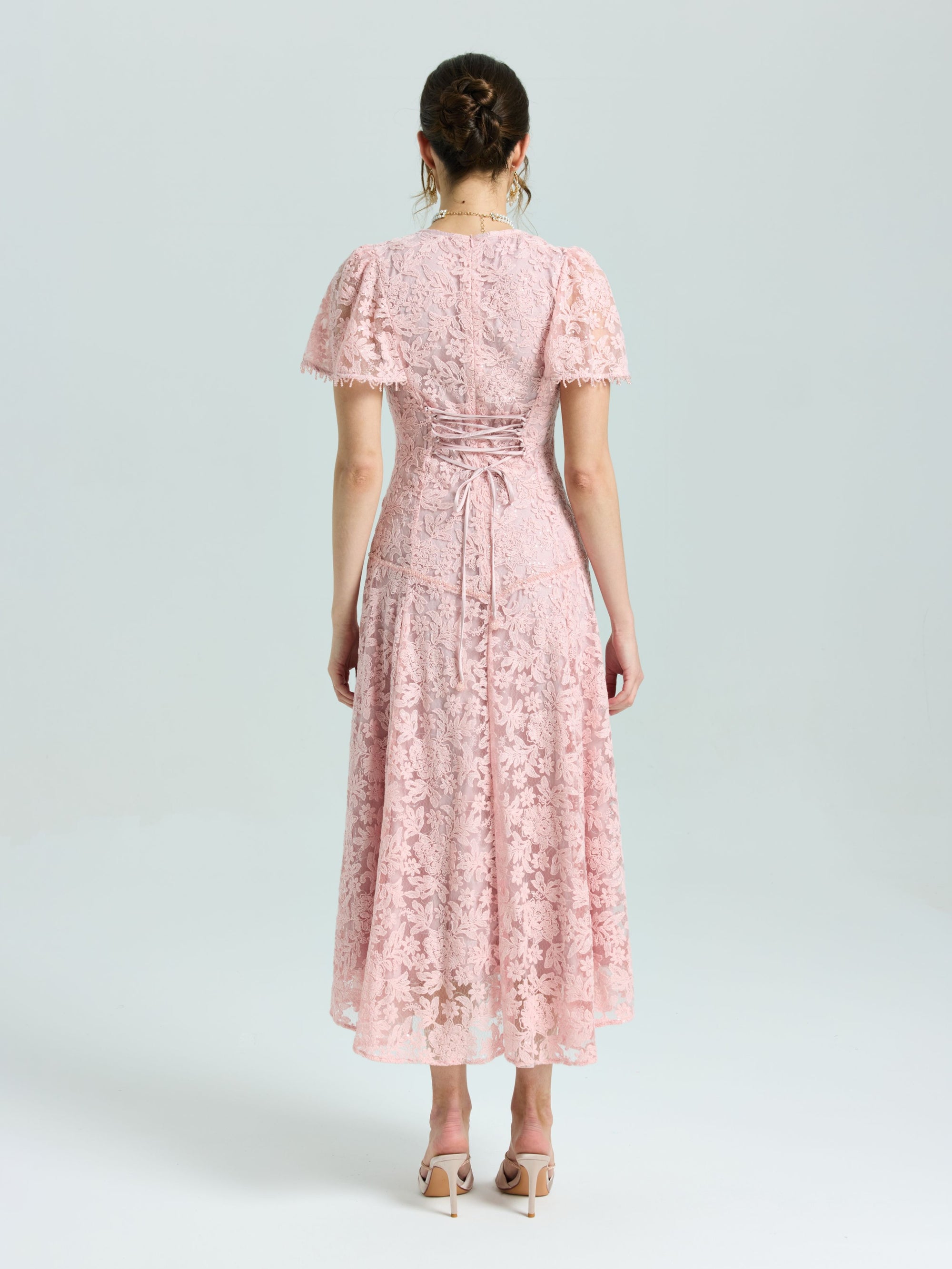 A woman faces away, wearing the Social Queen Floral Embroidered Lace Dress in light pink with short sleeves and lace-up back detail, paired with nude heeled sandals.