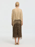 A person stands facing away, wearing a textured beige top with fringe, a brown sequined skirt, gold high-heeled sandals, and the Pearl Floral Appliqué Fringe Cape by Social Queen. Hair is styled in a low bun for an elegant touch.