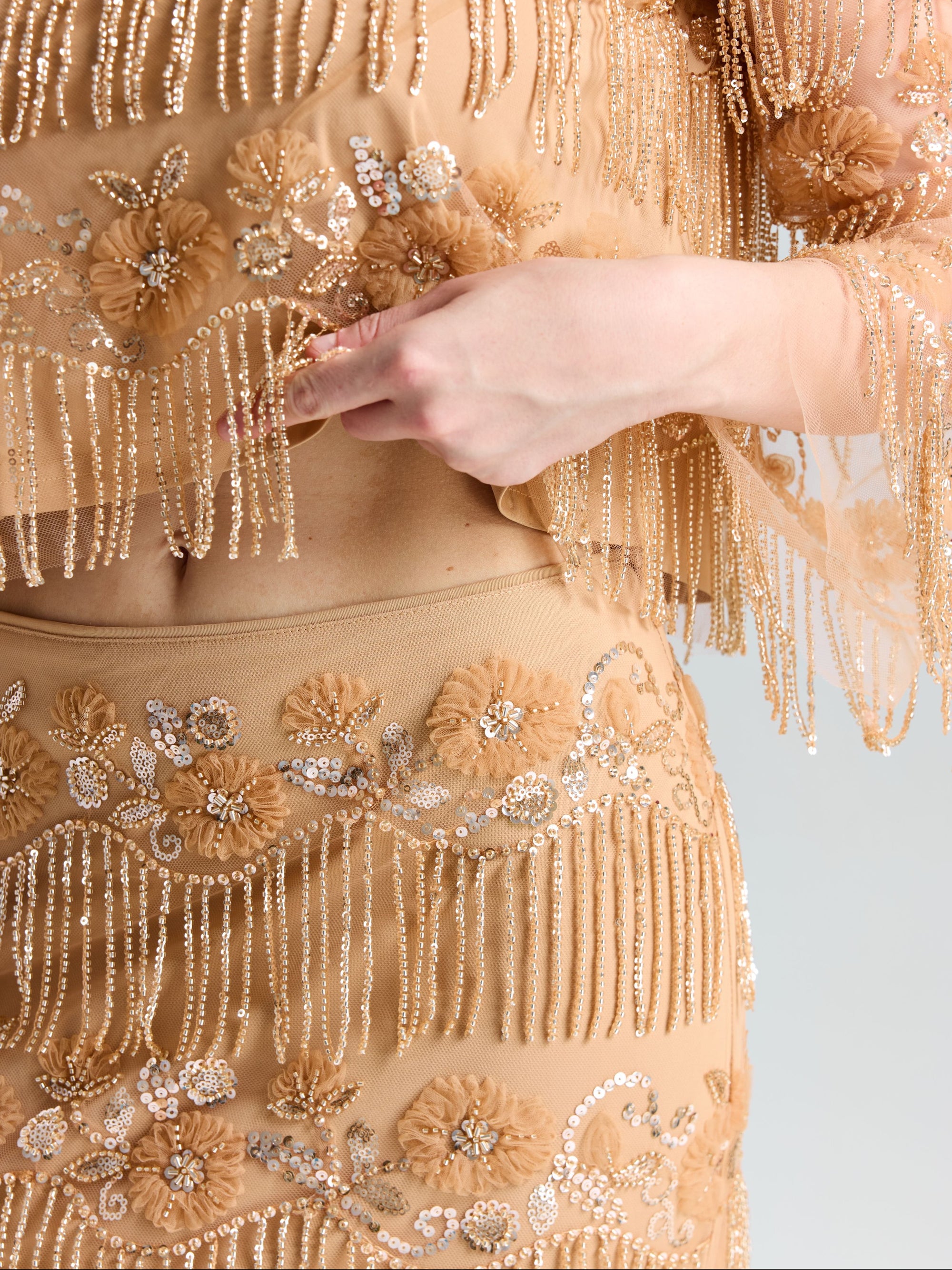 A person wearing the Social Queen Beaded Fringe Slit Midi Skirt, featuring beige floral embroidery, sequins, and beaded fringe, holds a section of the fabric with their right hand.