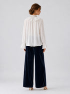 A woman stands facing away, wearing a white long-sleeve blouse with a ruffled collar and cuffs, paired with Social Queen’s Silk Velvet Wide-Leg Pants in navy crinkled texture and white heels, set against a plain light background.