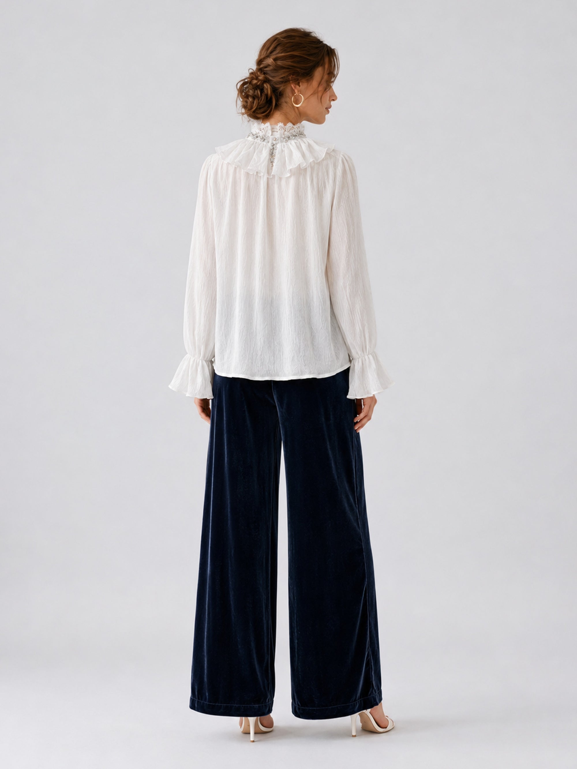 A woman stands facing away, wearing a white long-sleeve blouse with a ruffled collar and cuffs, paired with Social Queen’s Silk Velvet Wide-Leg Pants in navy crinkled texture and white heels, set against a plain light background.