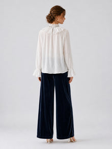 A woman stands facing away, wearing a white long-sleeve blouse with a ruffled collar and cuffs, paired with Social Queen’s Silk Velvet Wide-Leg Pants in navy crinkled texture and white heels, set against a plain light background.